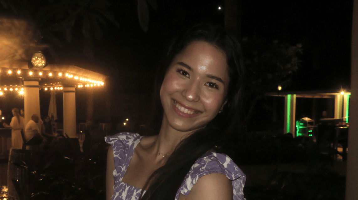 Young woman smiling with long dark hair, wearing a purple patterned top with ruffled sleeves, standing outdoors at night with warm lights and people in the background.